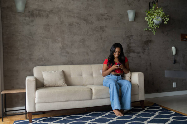 Young black woman working at home with smartphone sitting on her couch in the living room.  Home office concept. High quality photo