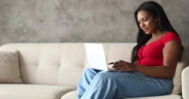 Genç siyahi kadın evde dizüstü bilgisayarıyla çalışıyor oturma odasında kanepesinde oturuyor. Merkez ofis konsepti. Çalışmak için gri defter. Yüksek kaliteli video