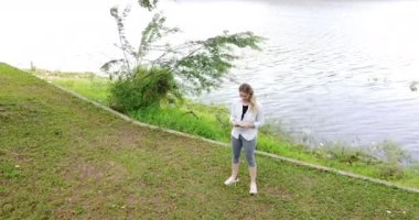 Gündüz vakti arka planda yeşil park olan akıllı telefon kullanan genç beyaz bir kadın. Su, göl. Yüksek kaliteli video. Cep telefonu, teknoloji, şehir kavramı.