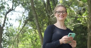 Gündüz vakti arka planda yeşil park olan akıllı telefon kullanan genç beyaz bir kadın. Yüksek kaliteli video. Cep telefonu, teknoloji, şehir kavramı.