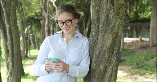 年轻的白人妇女在白天使用智能手机 背后有一个绿色的公园 高质量的视频 移动电话 城市概念 — 图库视频影像