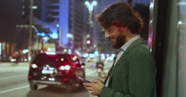 Arka planda şehir manzarası olan gece akıllı telefon kullanan genç bir adamın görüntüsü.