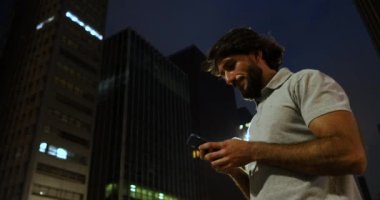 Arka planda şehir manzarası olan gece akıllı telefon kullanan genç bir adamın görüntüsü.