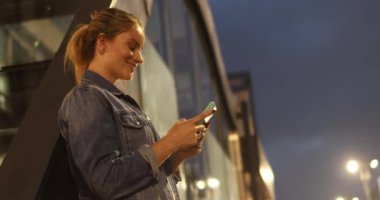 Gece vakti akıllı telefon kullanan genç bir kadın. Arka planda şehir manzarası var.. 