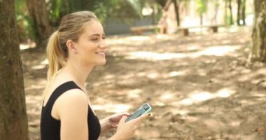 Gündüz vakti yeşil parklı akıllı telefon kullanan genç bir kadın. Yüksek kaliteli video. Cep telefonu, teknoloji, şehir kavramı.