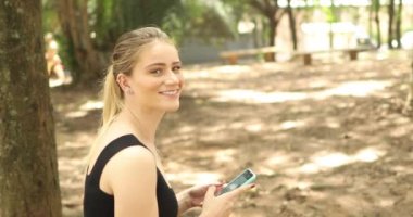 Gündüz vakti yeşil parklı akıllı telefon kullanan genç bir kadın. Yüksek kaliteli video. Cep telefonu, teknoloji, şehir kavramı.