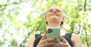 Gündüz vakti yeşil parklı akıllı telefon kullanan genç bir kadın. Yüksek kaliteli video. Cep telefonu, teknoloji, şehir kavramı.
