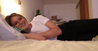 Genç bir kadın gece yatak odasındaki dairesinde akıllı telefon kullanarak yatağına uzanıyor. Yüksek kaliteli video. Cep telefonu, teknoloji, konsept.