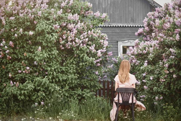Kalın yapraklı leylak çalılarının arasına saklanmış eski bir köy evinin arka planında, parlak bir bahar gününde, eski bir sandalyede oturan bir kız.