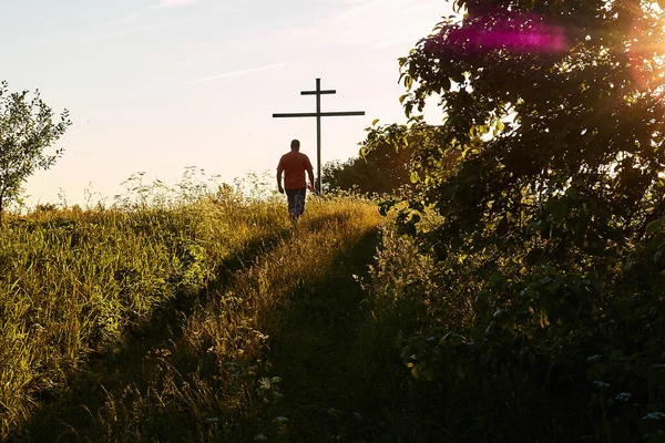 Ahşap bir haça doğru yürüyen bir adam, batan güneşin parlak altın renklerinde, giden yaz gününde.