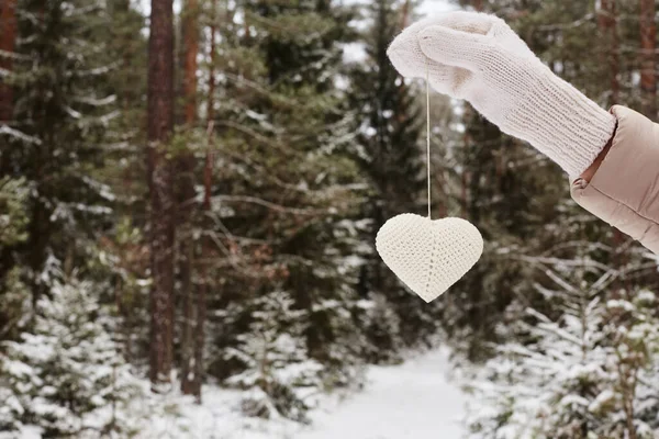 Beyaz ipliklerle işlenmiş bir kalp, yakın plan, bir kızın uzanan eline asılmış, kış ormanının bulanık arka planına karşı