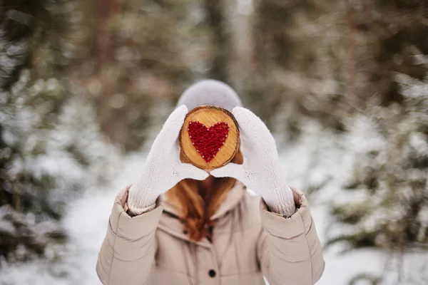 Kırmızı ipliklerle işlenmiş bir kalp, bir ağaç dalının kesiğinin ahşap bir tabanı üzerinde, kızın yüzünü kaplayan, kışın bulanık arka planına karşı