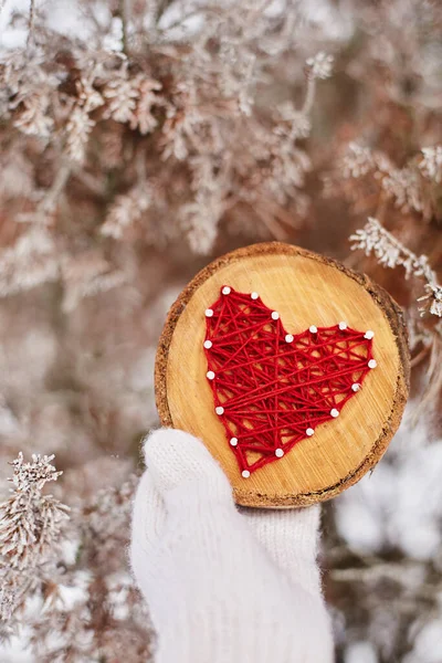 El yapımı bir kalp, kırmızı ipliklerle işlenmiş, bir ağaç dalının ahşap bir tabanında, beyaz eldivenli bir kadının elinde, karla kaplı bir ormanın bulanık arka planında