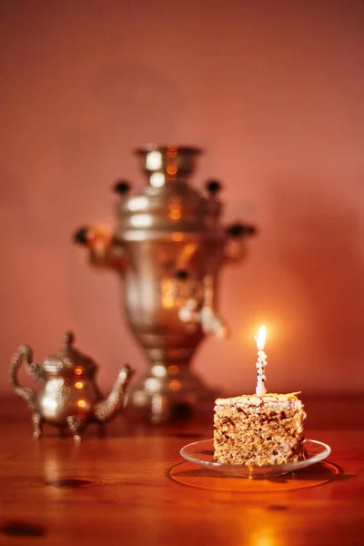 Tabağında mum, arka planında semaver ve çaydanlık duran bir kek parçasının şenlikli natürmortu. Bulanık odaklı arkaplan