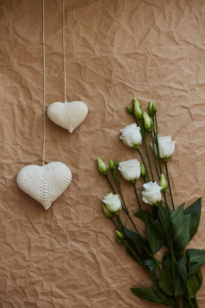 Örgü örülmüş beyaz kalpler ve bir buket beyaz Eustoma çiçekleri tek bir açık kahverengi zemin üzerinde buruşmuş kağıt dokusu ile