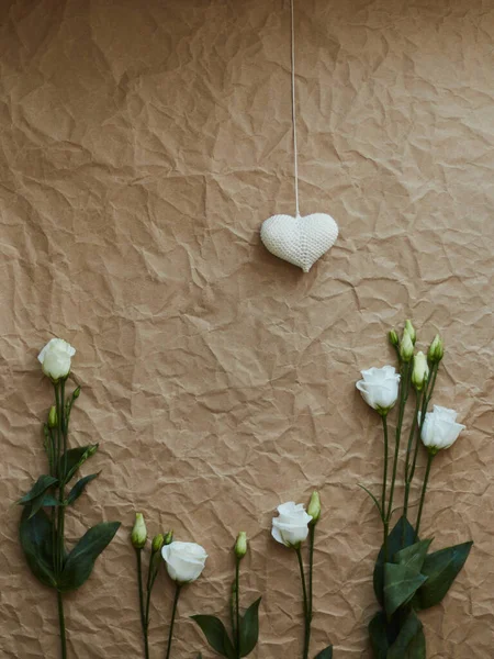 Eşit bir şekilde yerleştirilmiş, beyaz östoma çiçeklerinden oluşan şenlikli bir kompozisyon ve buruşuk kağıt dokusuna sahip tek bir açık kahverengi arka planda hafif iplikten örülmüş bir kalp.