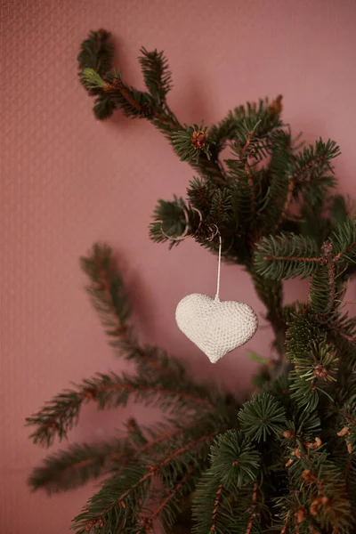 Taze bir köknar dalına asılmış, açık pembe arka planda beyaz bir kalp.