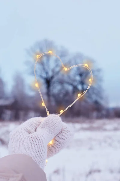 Decorative heart in a woman\'s hand, dressed in a white mitt, made of bright lights, on a white snowy background