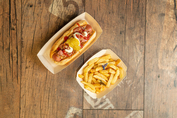 Вид сверху на хот-дог с хрустящим луком, вареной колбасой, жареным беконом, майонезом, горчицей и кетчупом, украшенный картошкой фри в пакетах для выноса