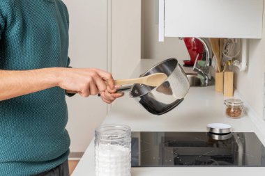 Genç adam siyah seramik bir hob üzerinde ahşap kaşıkla paslanmaz çelik bir tavayla yemek pişiriyor.