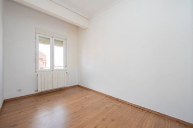 Beyaz boyalı duvarları ve alüminyum kaloriferli kestane döşemesi olan boş bir yatak odası.