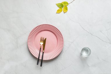Çatal bıçaklı pembe tabak, ağaç yaprakları, mermer masa, tatlı suyla kristal bardak.