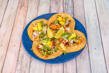 Mavi metal bir tabak, içinde taco al pastor ve et, soğan, mango, limon ve kişniş var.