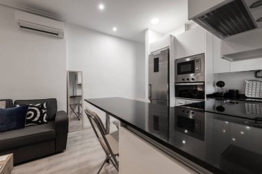 Paslanmaz çelik aletli mutfak, siyah mermer adalı beyaz dolaplar ve karşı tarafında kanepe olan oturma odası.