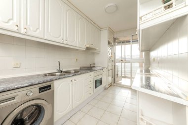 Beyaz ahşap mobilyalı uzun mutfak, mavi tezgah ve terasa erişimi olan aletler.