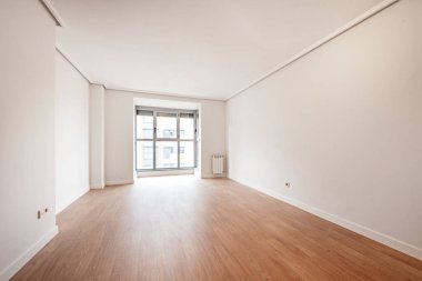 Arka planda büyük penceresi olan boş bir oturma odası, ahşap zemin ve beyaz boyalı duvarlar.