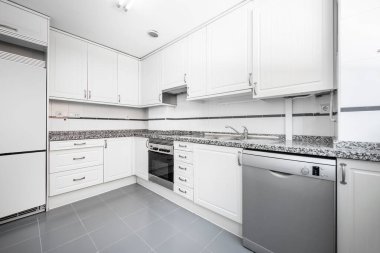 Duvarları beyaz ahşap dolaplarla kaplı mutfak, gri granit tezgah, gri bulaşık makinesi, gri porselen döşeme ve uygun vale.