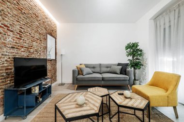 Gri kanepesi, televizyonu, yuvarlak cam yemek masası, beyaz perdeleri, sarı kişisel koltukları, mavi metal TV masası, düşük çokyüzlü masaları ve korunmasız tuğla duvarı olan oturma odası.