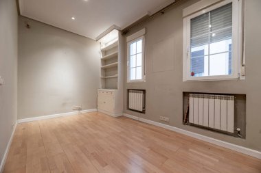 Duvarda alçı ve ahşap mobilyalar, tahta döşeme, beyaz alüminyum kalorifer ve beyaz alüminyum pencereli oturma odasının önü.