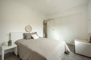 Büyük boy yatak odası, gri yatak örtüsü, yastıklar ve yatak başlığının üstündeki yuvarlak ayna ve gri parke döşeme.