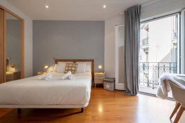 Büyük yataklı yatak odası, ahşap çalışma masası, ahşap döşeme ve ayna kapılı gardırop ve çift pencereli balkon.