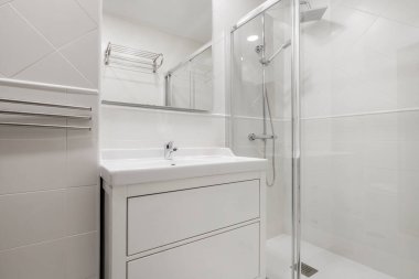 Cam ve çelik duşlu küçük beyaz banyo, kare çerçevesiz ayna ve beyaz porselen lavabo.