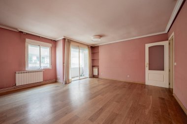 Terasa erişimi olan boş bir oturma odası, alüminyum radyatörler, pembe boyalı duvarlar ve ahşap döşemeler.