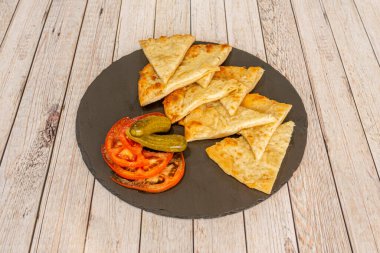 Dilimlenmiş peynirli omlet, domates dilimleri ve turşuları açık ahşap bir masada siyah bir tabakta.