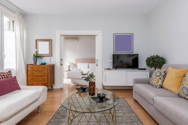 Kanepeleri kumaşla döşenmiş oturma odası, düz ekran televizyonlu ahşap büfe, yuvarlak cam sehpa ve arka plandaki yatak odasına giriş.