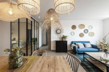 Ahşap mobilyaları ve mavi kumaş koltuğu olan şık dekorasyonlu oturma odası, hasır lambalar ve cam bölme ve siyah metal.