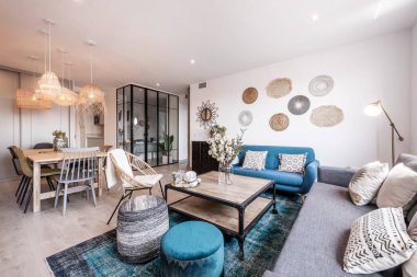 Oturma odası ahşap yemek masası döşenmiş ahşap sandalyeler, hasır lambalar, hafif ahşap zemin ve halı ve siyah metal ve cam bölmeli mavi kanepe.