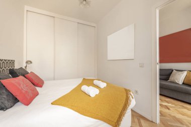 Büyük boy yataklı yatak odası, beyaz sürgülü kapılı gardırop, kırmızı ve siyah yastıklar ve hardal renkli battaniye.