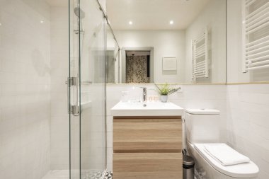 Ahşap mobilyaların üzerine asılmış porselen lavabo, duvardan duvara ayna, beyaz ısıtılmış havlu korkuluğu, krom aksesuarlar, sürgülü cam ekranlı duş kabini.