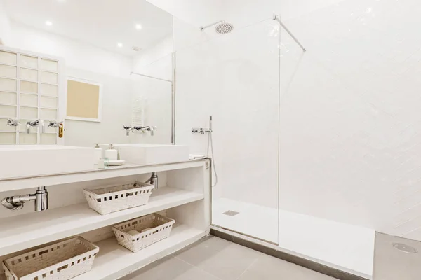 Bathroom with built-in vanity unit with white porcelain sink, wicker baskets, frameless mirror and glass-enclosed shower stall with vintage chrome taps