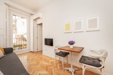 Demir ayaklı ahşap masalı oturma odası, beyaz tahta sandalyeler, siyah metal parmaklıklı balkon ve şehrin çatılarının manzarası.