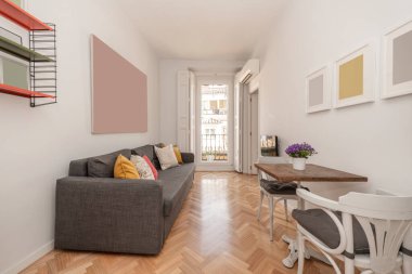 İki kişi için ahşap ve metal masa, tahta sandalyeler, bir sürü yastıklı gri kanepe ve renkli raflar, beyaz ahşap panjurlu balkon