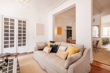 Üç koltuklu, kumaşla döşenmiş büyük bir oturma odası. Yastıklar, ahşap döşemeler, metal bir sehpa ve aynalar ve kapılarda ahşap marangozluk..