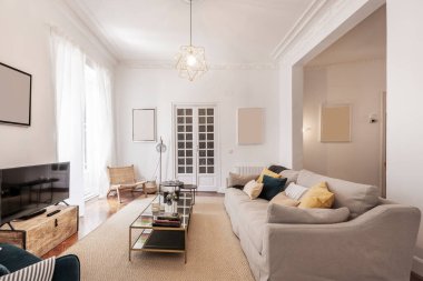 Üç koltuklu bir oturma odası, kumaşla döşenmiş, bir sürü yastıklı, metal bir sehpa ve aynalar, ve kapılarda ahşap kaplama, yerde açık renkli bir halı ve bir duvarda televizyon.