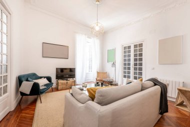 Üç koltuklu oturma odası, kumaşla döşenmiş bir sürü yastık, beyaz kapılı balkon ve kapılarında marangozluk, yerde hafif halı ve bir duvarda televizyon.