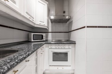 Siyah kuvars görünümlü tezgahı olan bir mutfağın köşesi ve beyaz mikrodalga fırını olan beyaz dolaplar, paslanmaz çelik kaput ve kenarlı beyaz fayanslar.